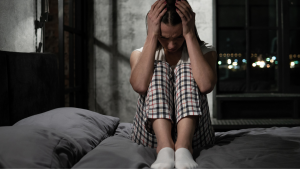 Eine Frau sitzt nachts wach im Bett, den Kopf auf die Hände gestützt, mit gerunzelter Stirn, und kann wegen kreisender Gedanken nicht einschlafen.